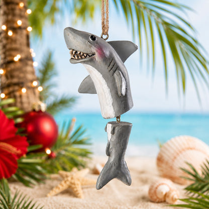 A shark Christmas ornament hanging from a rope, styled on a beach-themed background with palm leaves, shells, sand, and holiday decorations.