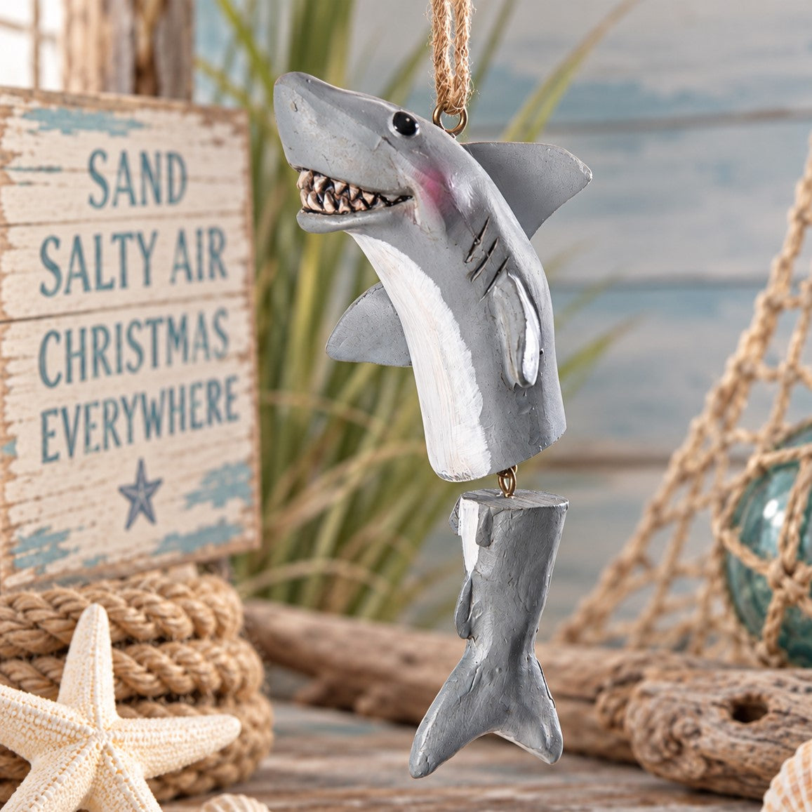 A gray and white shark Christmas ornament hanging from a rope, styled with nautical props including a starfish, rope, a wooden sign reading 'Sand Salty Air Christmas Everywhere,' and a blurred beach-themed background.