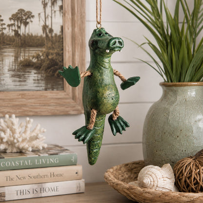 A green alligator hanging ornament with rope limbs and a rope hanger, displayed in a coastal-style home decor setting next to a potted plant and books. Collectible ornament by Bert Anderson.