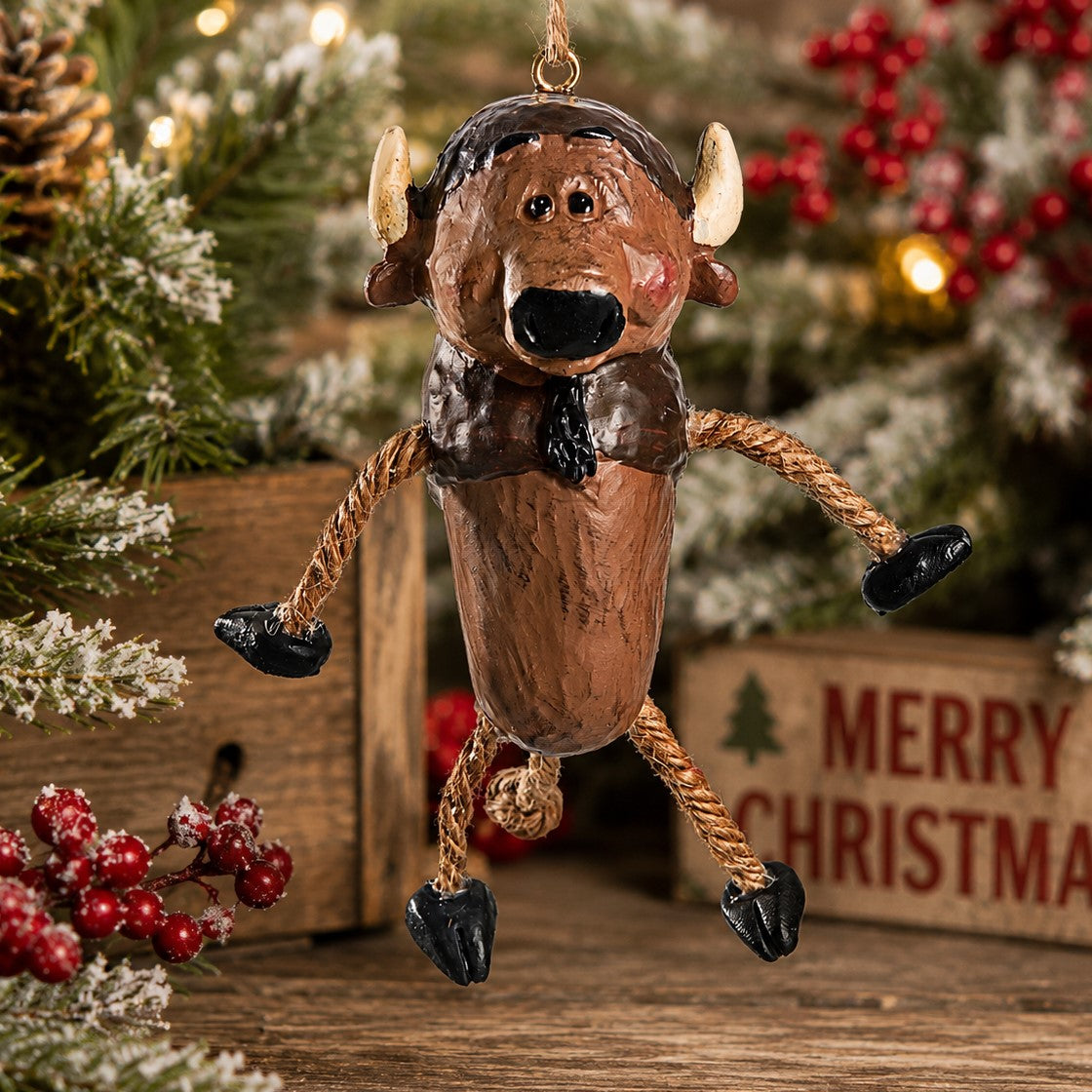 A rustic Buffalo Christmas ornament with rope legs, black hooves, a dark brown hat and bow tie, and small cream horns. The ornament is hanging in front of a blurred holiday tree and a wooden sign that reads Merry Christmas.  Designed by artist Bert Anderosn.