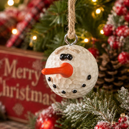 A Christmas ornament resembing a golf ball with a carved face, black bead eyes and buttons, and an orange carrot nose, hanging on a decorated Christmas tree with a blurred 'Merry Christmas' sign in the background.