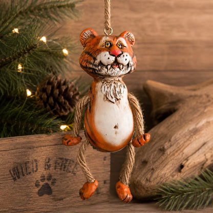 A whimsical tiger hanging ornament made from a metal tin with a painted orange and white body, hanging from a rope loop. The ornament is styled on a rustic wooden surface with pine branches, a pinecone, and driftwood in the background.