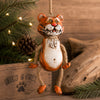 A whimsical tiger hanging ornament made from a metal tin with a painted orange and white body, hanging from a rope loop. The ornament is styled on a rustic wooden surface with pine branches, a pinecone, and driftwood in the background.