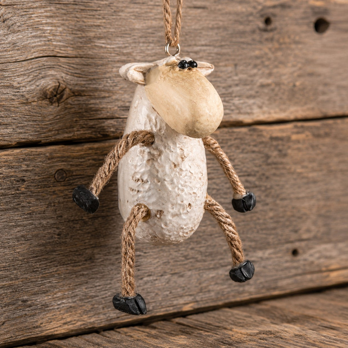 A rustic sheep-shaped hanging ornament made from textured cream-colored resin with rope legs and black hooves, shown against a weathered wooden plank background.