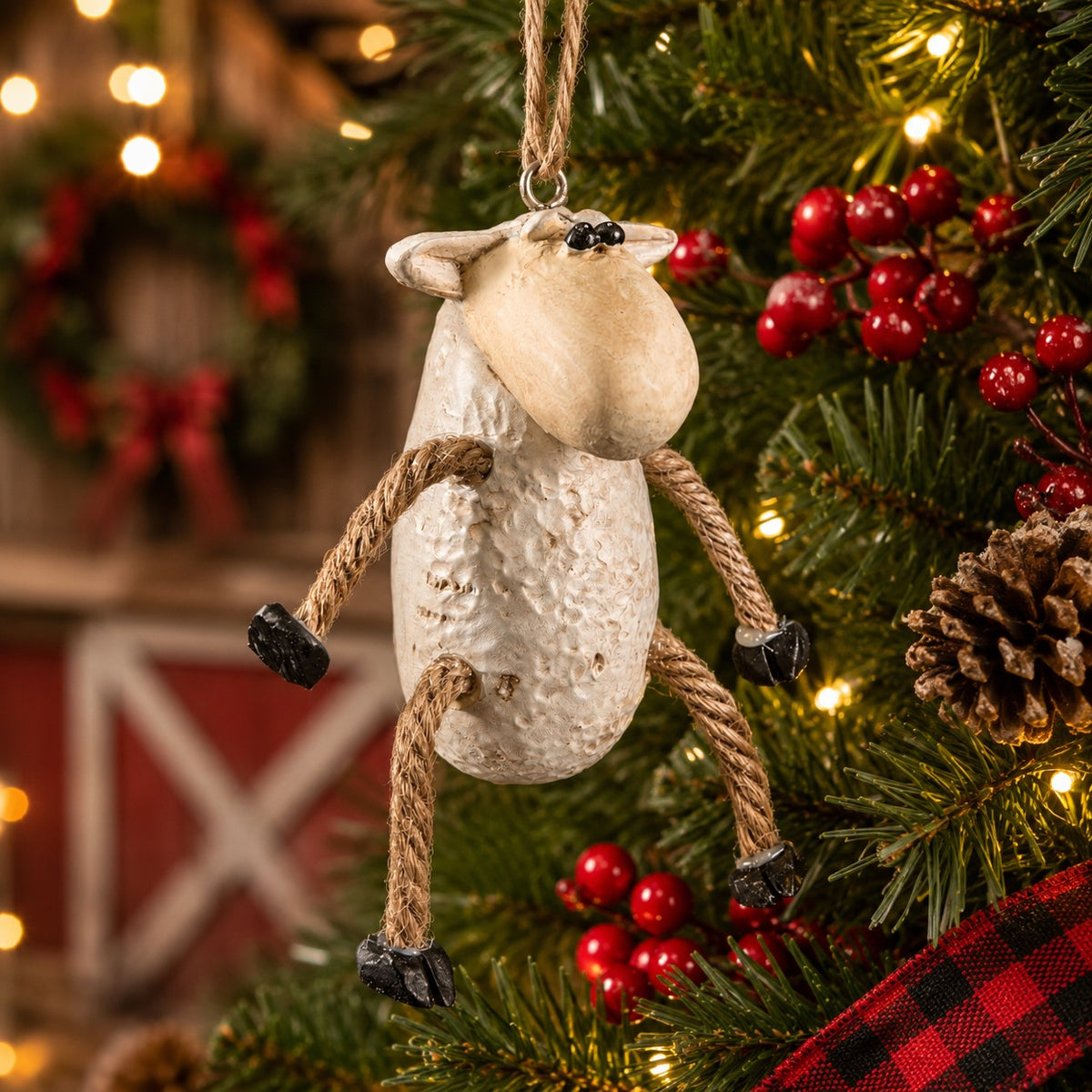 A rustic hanging sheep ornament made from a textured cream-colored resin or stone-like material, with rope legs and black hooves, hanging on a decorated Christmas tree with red berries, pinecones, and warm string lights.
