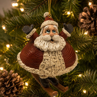 A Santa Claus hanging ornament on a Christmas tree, with a gold hanging loop, red coat, cream beard and trim, black mittens, and wooden-style skis, photographed among green tree branches and warm white lights.