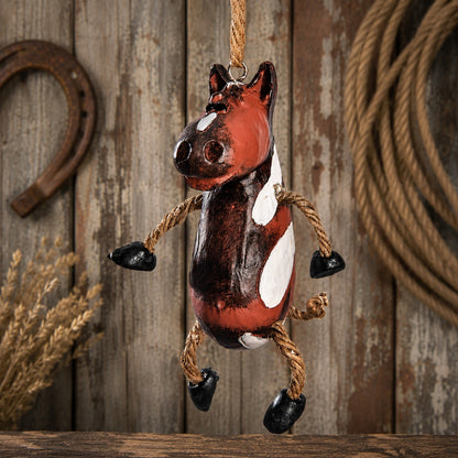 A rustic hanging horse painted in red, brown, black, and white with rope limbs and hooves, suspended against a weathered wood plank background with a horseshoe and rope props.
