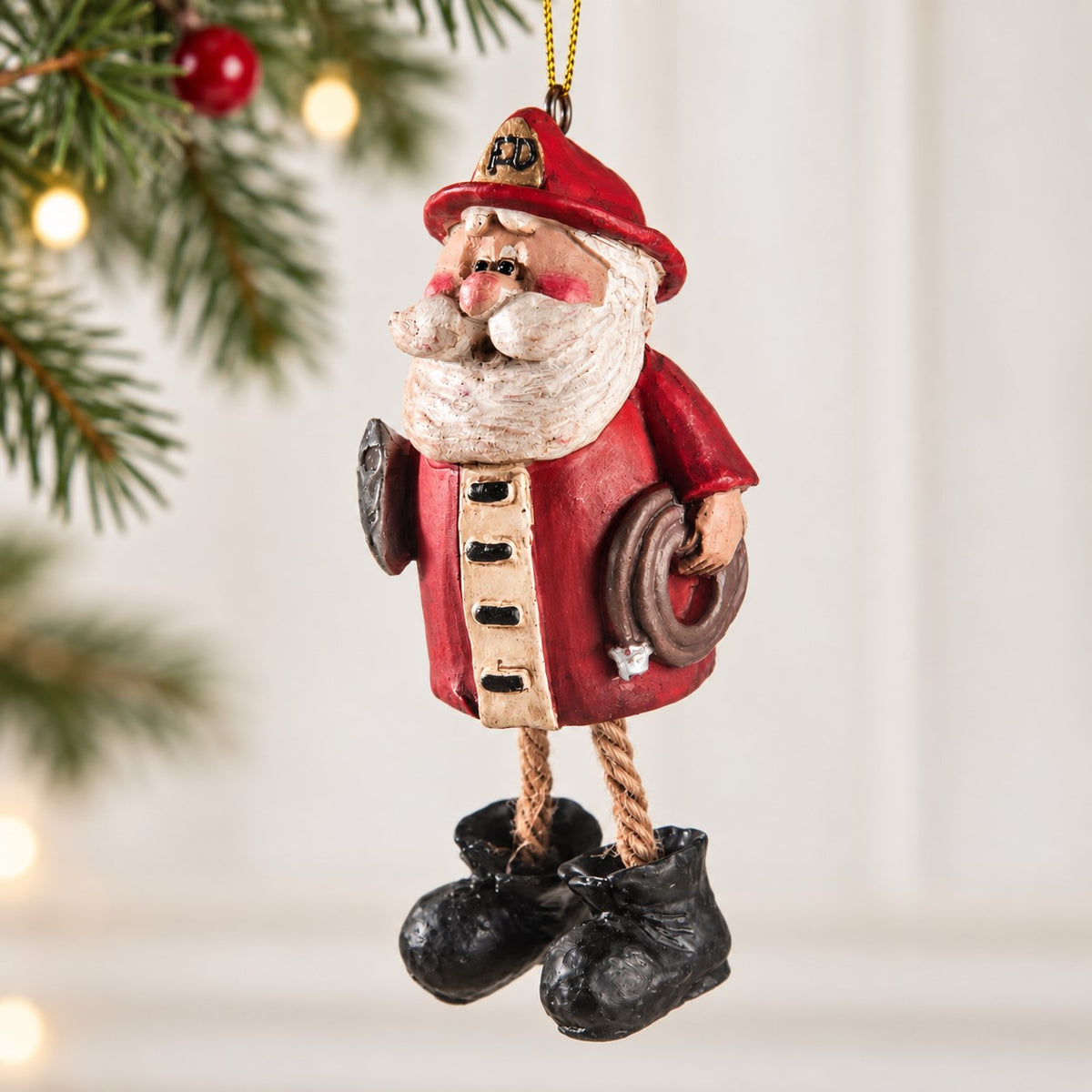 Bert Anderson firefighter Santa ornament with red fire helmet, fire hose, dangling rope legs and black boots in a bright Christmas setting.