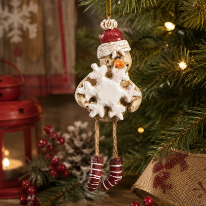A rustic snowman Christmas ornament hanging from a decorated tree. Ready for your winter deccor this snowman   is carrying a white snowflake and has a red and white knit hat, and red-and-white striped socks.