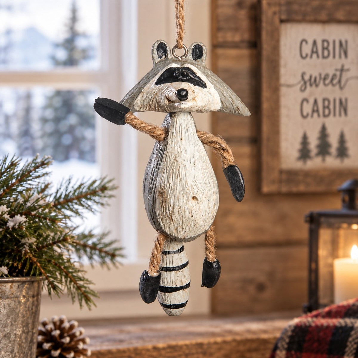 Bert Anderson raccoon ornament with dangling arms and legs displayed in a cozy rustic cabin setting.