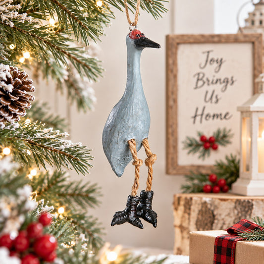 Bert Anderson Sandhill Crane ornament with dangling legs displayed in bright Christmas home décor with greenery, warm lights and red berries.