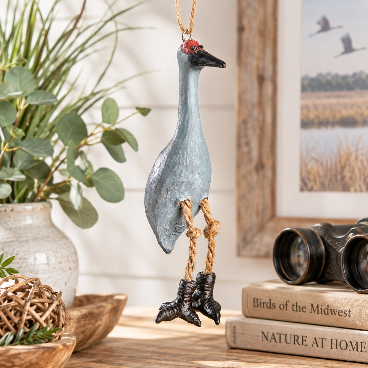 Bert Anderson Sandhill Crane ornament with dangling legs displayed in light nature-inspired home décor with binoculars and bird artwork.