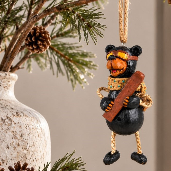 A black bear Christmas ornament wearing yellow goggles and holding a skis, hanging  in from a pine branch.