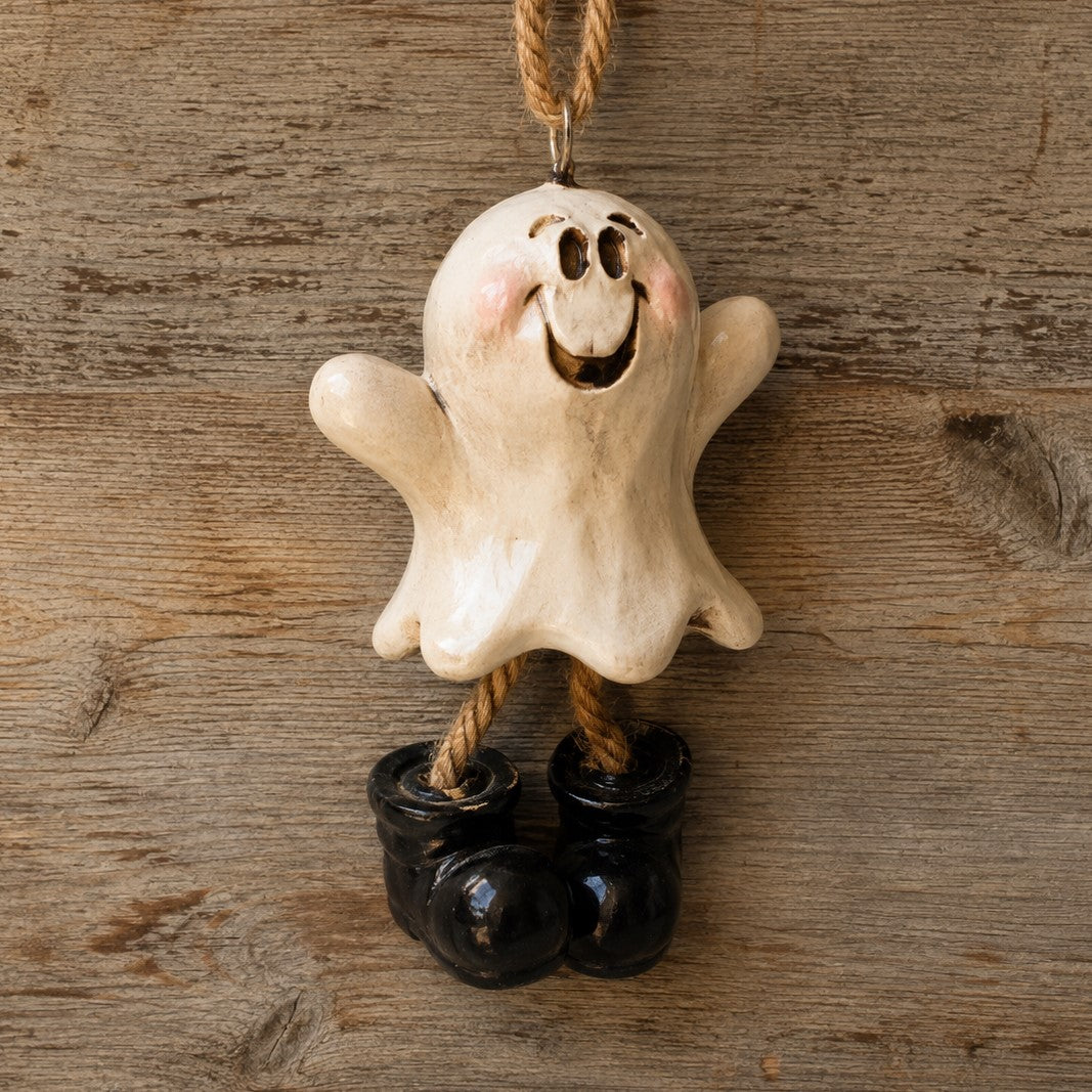 A ceramic Halloween ghost ornament with a cream-colored body, black boots, and a hanging loop, shown against a rustic wooden background.