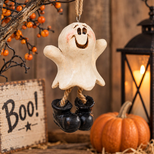 Bert Anderson ghost ornament with dangling black boots displayed in rustic Halloween décor with a pumpkin, lantern and orange berry branches.