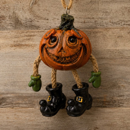 A Halloween hanging ornament shaped like a carved orange pumpkin with a smiling face, rope arms, green gloves, and black witch boots, displayed against a rustic wood plank background.