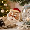 A festive Santa Claus Christmas ornament hanging from a pine branch, styled on a rustic tabletop with a blurred Christmas decor.