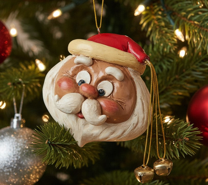 A festive Santa Claus Christmas ornament hanging on a decorated Christmas tree. The ornament features a detailed Santa face with a red hat, gold trim, white beard, and rosy cheeks, with small gold bells dangling from the side.