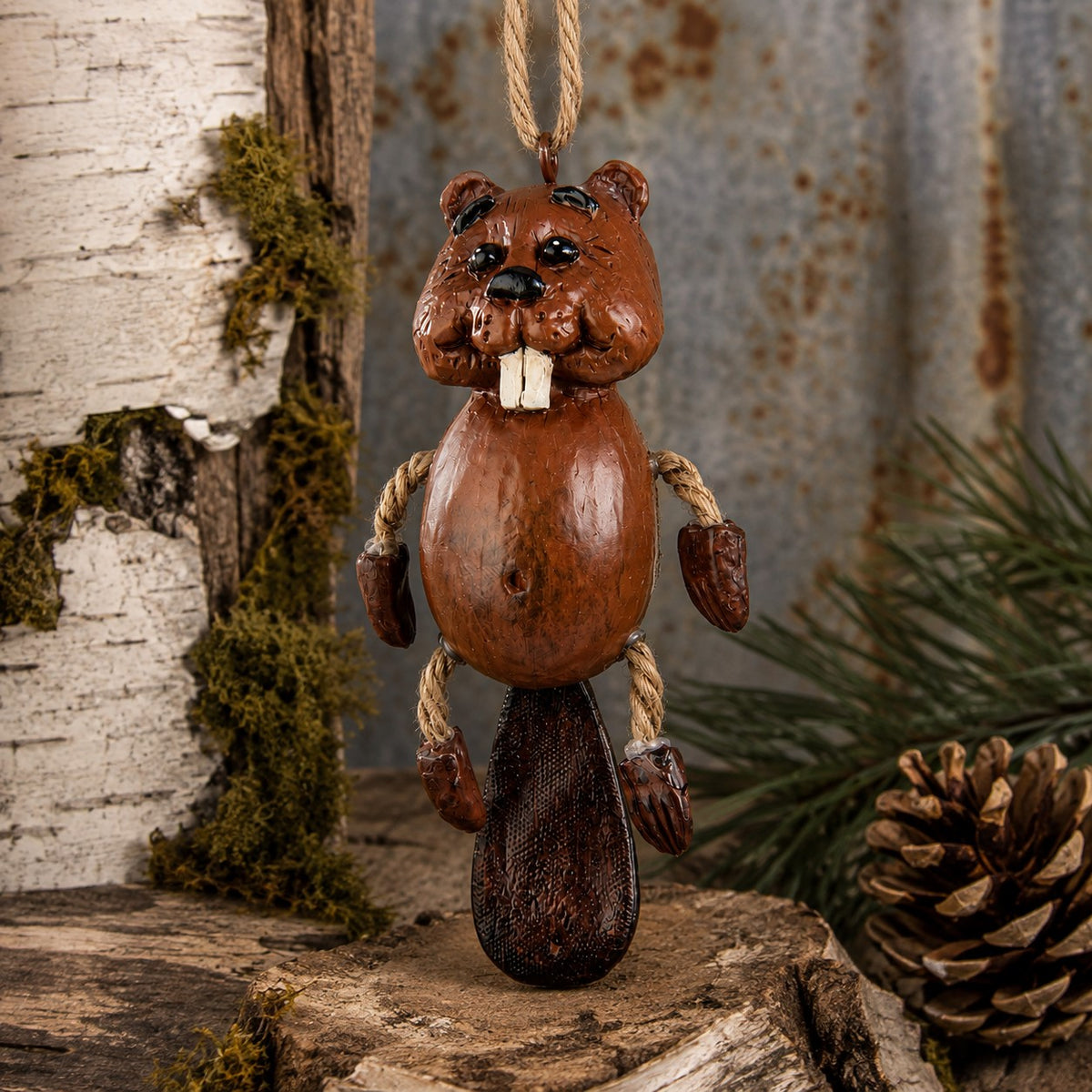 A whimsical beaver ornament with rope limbs and a hanging loop, displayed on a rustic log with birch bark, moss, pine needles, and a pinecone in the background.