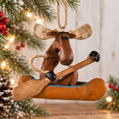 Bert Anderson moose canoe ornament with paddle hanging among snowy evergreen branches, red berries and warm Christmas lights.