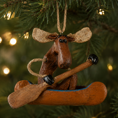 A rustic moose ornament in a canoe hanging on a Christmas tree, with pine branches and warm string lights in the background.