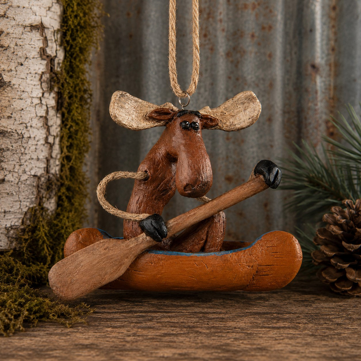 A rustic moose-shaped hanging ornament sitting in a small canoe with a wooden paddle, styled on a wood surface with bark, moss, pine needles, and a pinecone in the background.