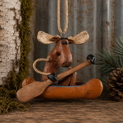 A rustic moose-shaped hanging ornament sitting in a small canoe with a wooden paddle, styled on a wood surface with bark, moss, pine needles, and a pinecone in the background.