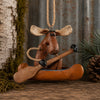 A rustic moose-shaped hanging ornament sitting in a small canoe with a wooden paddle, styled on a wood surface with bark, moss, pine needles, and a pinecone in the background.
