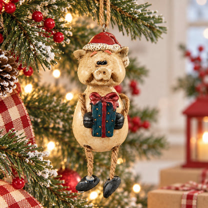 Bert Anderson pig Christmas ornament wearing a Santa hat and holding a wrapped gift, hanging among snowy evergreen branches and warm holiday lights.