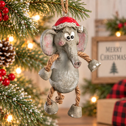 Bert Anderson elephant Christmas ornament wearing a red Santa hat with dangling rope arms and legs, hanging among snowy evergreen branches and warm holiday lights.