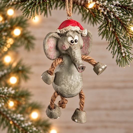 Bert Anderson elephant Christmas ornament wearing a Santa hat with dangly rope legs, displayed with evergreen branches and warm holiday lights.