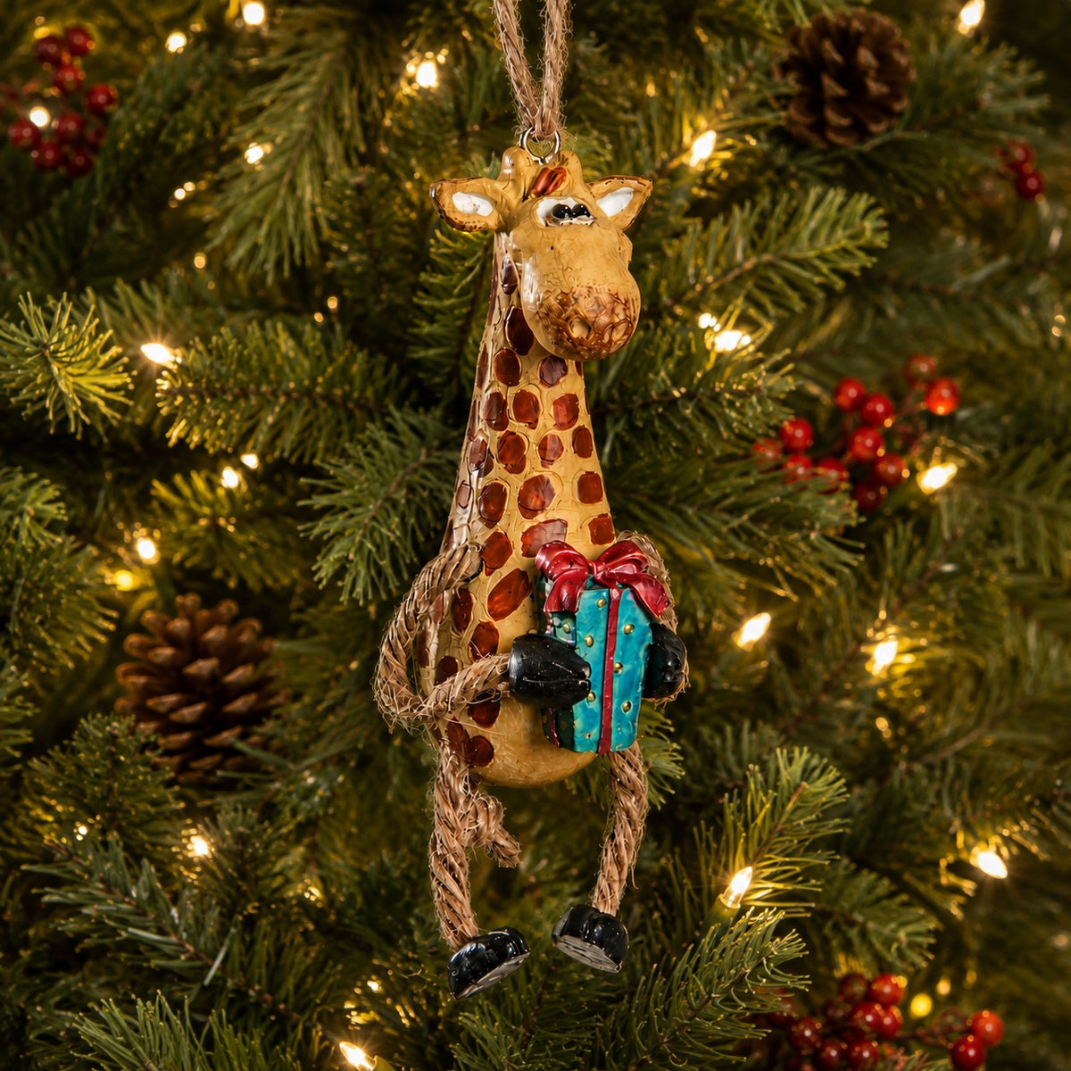 A giraffe Christmas ornament hanging on a lit green Christmas tree, holding a blue gift box with a red bow.