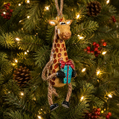 A giraffe Christmas ornament hanging on a lit green Christmas tree, holding a blue gift box with a red bow.