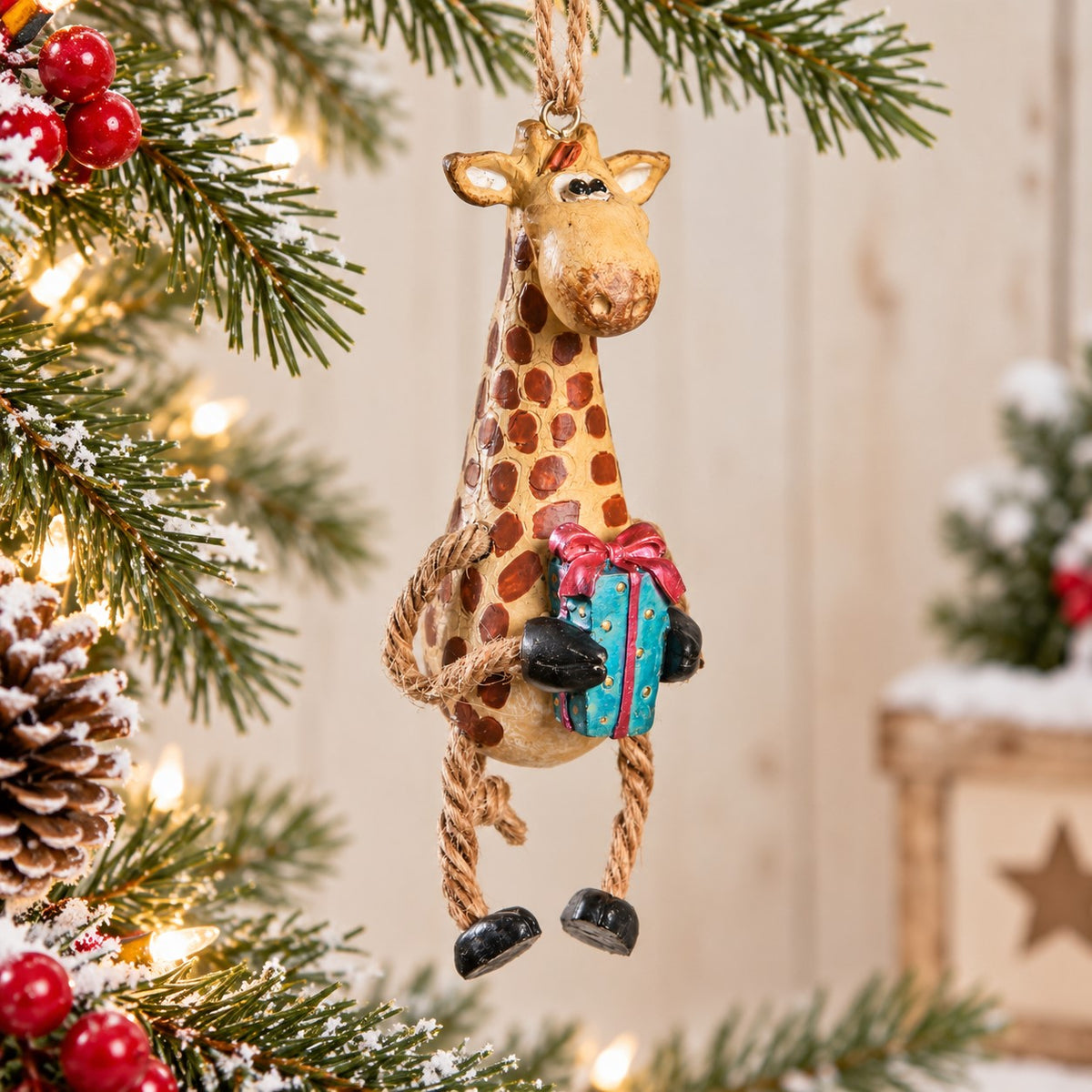 Bert Anderson giraffe ornament holding a colorful Christmas gift, hanging among snowy evergreen branches, red berries and warm holiday lights.