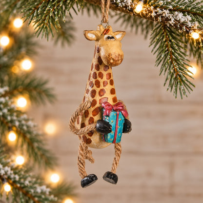 Bert Anderson giraffe Christmas ornament holding a colorful present with dangly rope legs, displayed with evergreen branches and warm holiday lights.