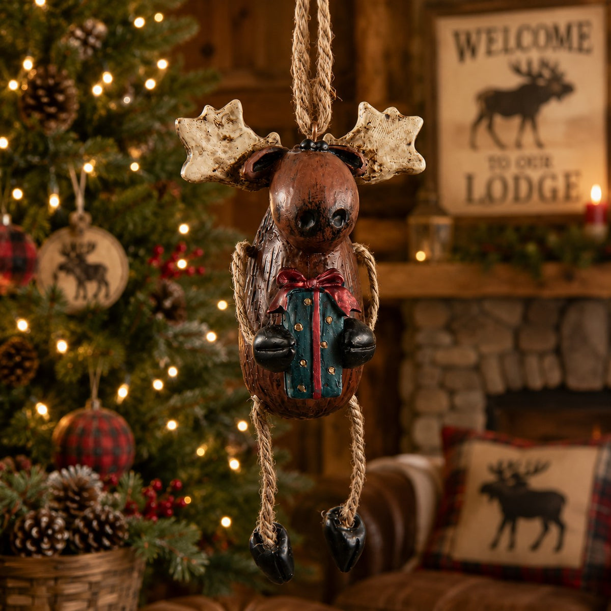 A rustic Christmas Moose ornament  hanging in front of a decorated Christmas tree and fireplace. The moose has antlers, a brown body, black feet, and holds a small wrapped gift with a red bow.