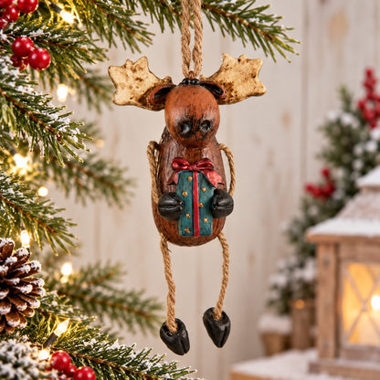 Bert Anderson moose ornament holding a Christmas gift, hanging among snowy evergreen branches, red berries, pinecones and warm holiday lights.