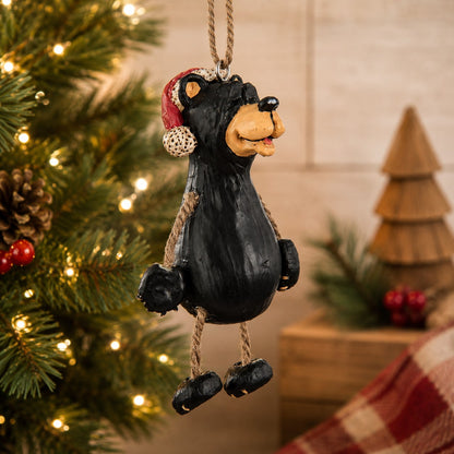 A black bear Christmas ornament  wearing a Santa hat hanging from a tree with warm string lights in the background.