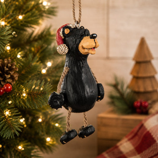 A black bear Christmas ornament  wearing a Santa hat hanging from a tree with warm string lights in the background.