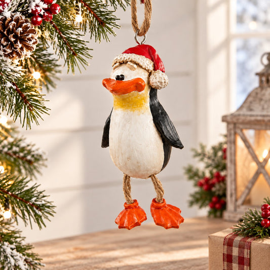 Bert Anderson penguin ornament with Santa hat and dangly orange feet displayed against a light, softly decorated Christmas background.