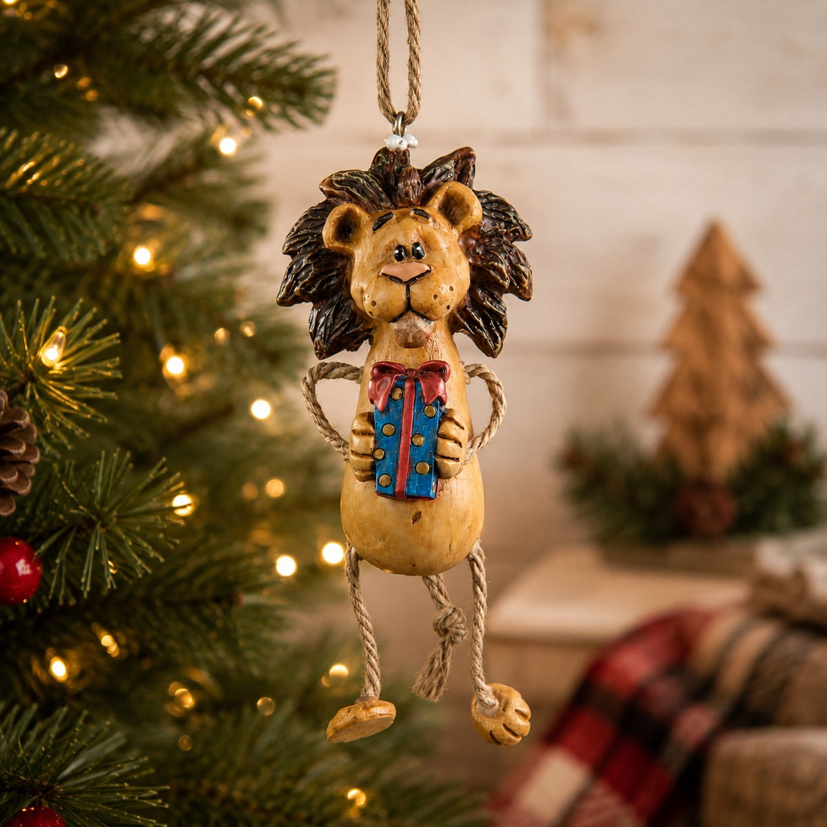 A hanging lion Christmas ornament  holding a blue gift box, displayed in front of a lit Christmas tree.