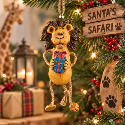 Bert Anderson African safari lion Christmas ornament holding a wrapped gift, hanging among festive greenery with safari-inspired holiday décor.