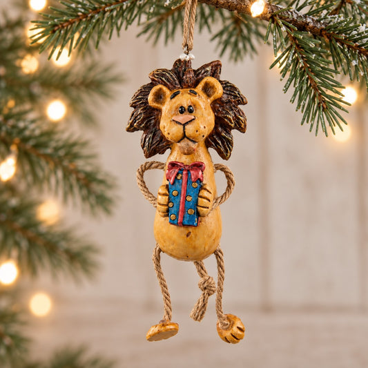 Bert Anderson lion Christmas ornament holding a colorful present with dangly rope legs, displayed with evergreen branches and warm holiday lights.