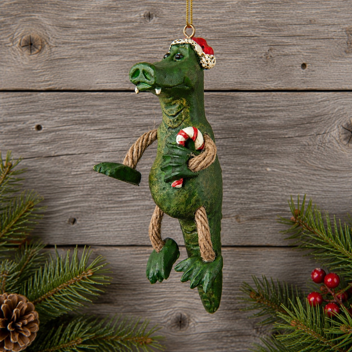 A green alligator Christmas ornament with rope limbs and a Santa hat, hanging against a rustic wood plank background with pine branches and red berries at the edges.