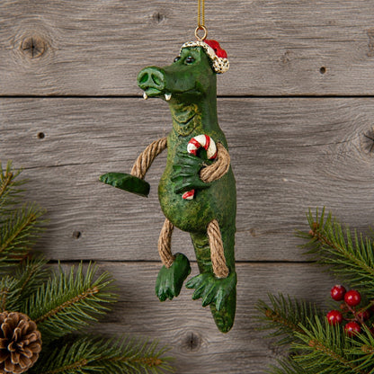 A green alligator Christmas ornament with rope limbs and a Santa hat, hanging against a rustic wood plank background with pine branches and red berries at the edges.