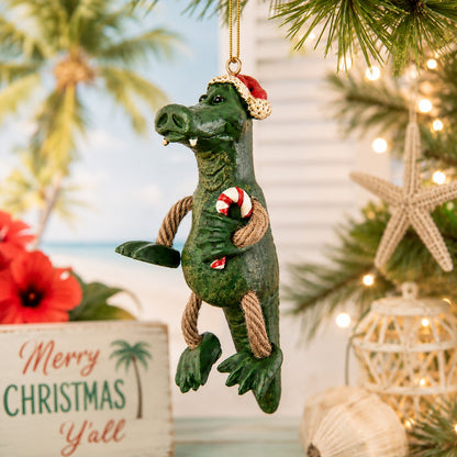 A green alligator Christmas ornament hanging from a pine tree, wearing a red Santa hat and holding a red-and-white candy cane. The background shows a softly blurred beach-themed holiday scene with palm trees, warm string lights, a starfish, and a sign reading 'Merry Christmas Y'all.