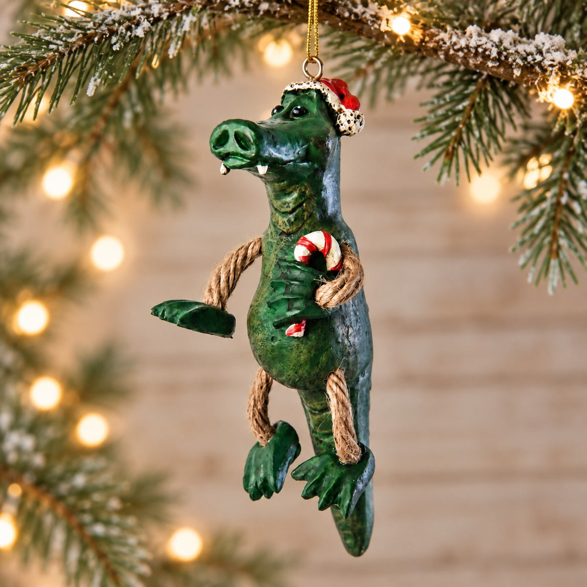 Bert Anderson alligator Christmas ornament wearing a Santa hat and holding a candy cane, displayed with evergreen branches and warm holiday lights.