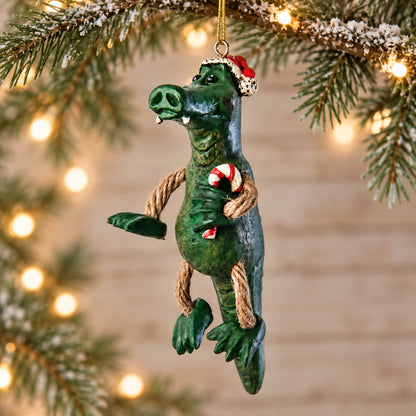 Bert Anderson alligator Christmas ornament wearing a Santa hat and holding a candy cane, displayed with evergreen branches and warm holiday lights.