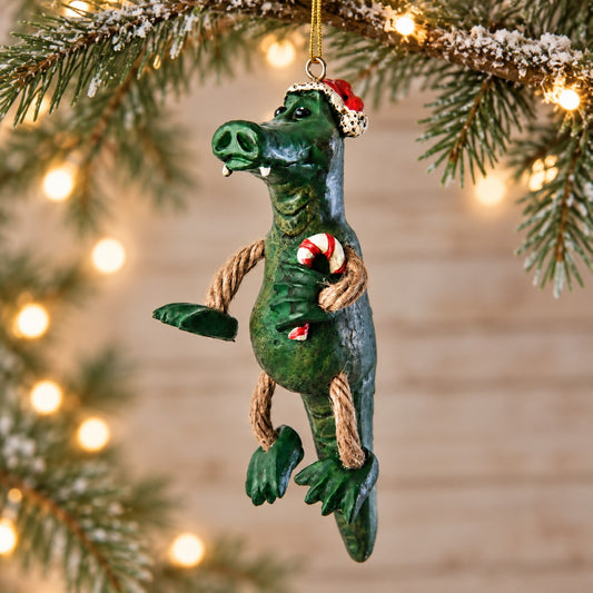 Bert Anderson alligator Christmas ornament wearing a Santa hat and holding a candy cane, displayed with evergreen branches and warm holiday lights.