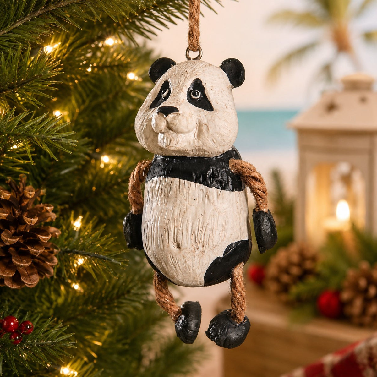 A rustic panda Christmas ornament hanging from a tree branch, with warm holiday lights and a softly blurred beach-themed background.